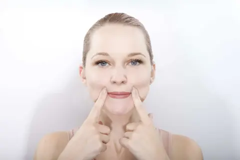 Une femme pratique le yoga du visage, ses doigts pressant ses joues pour un sourire accentué.