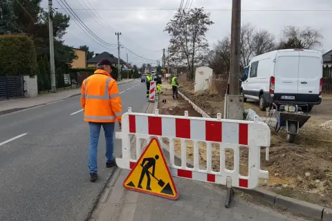 Robotnicy w kamizelkach odblaskowych pracują na budowie przy drodze, ogrodzonej barierkami.
