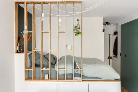 Un studio avec un coin nuit séparé par une étagère en bois. Le lit est fait de draps verts pâles.