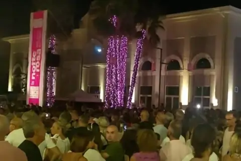 Folla festante a Porto Sant'Elpidio per la Notte Rosa, con palme illuminate di rosa e un grande striscione.