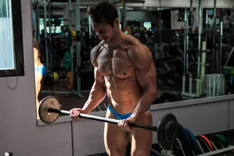 Atleta in palestra, con bicipite e tricipite scolpiti, solleva un bilanciere. Riflessi nello specchio mostrano attrezzature sportive.