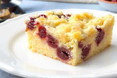 Ein Stück saftiger Kirschstreuselkuchen vom Blech auf einem weißen Teller, mit vielen Kirschen und knusprigen Streuseln.