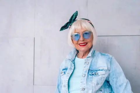 Modna starsza pani w okularach i jeansowej kurtce. Jej stylizacje, w tym spodnie z dziurami, są odważne i pełne życia.