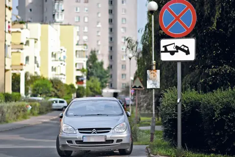 Srebrny samochód na drodze wewnętrznej, obok znaki zakazu zatrzymywania się i holowania. W tle bloki mieszkalne.