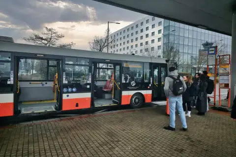 Ludzie czekają na autobus, który może być odpowiedzią na pytanie "jak dojechać do Pragi". W tle nowoczesny budynek i niebo.