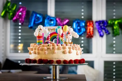 Kolorowy tort z jednorożcami i tęczą, idealny na zabawy na urodziny 7 latka. Balony i napis "Happy Birthday" w tle.