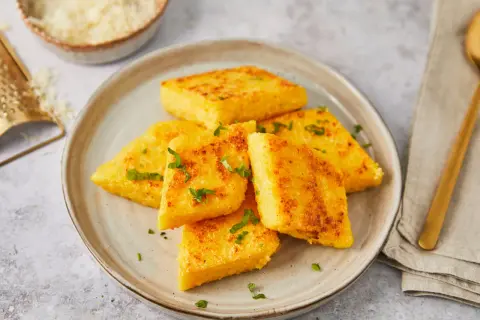 Gebratene Polenta-Ecken, italienisch garniert mit Petersilie, auf einem Teller. Daneben eine Reibe mit Parmesan.