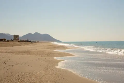 La playa de San Miguel, con sus aguas turquesas y arena dorada, se extiende hacia unas montañas. En la orilla, barcos y una torre antigua esperan la marea.