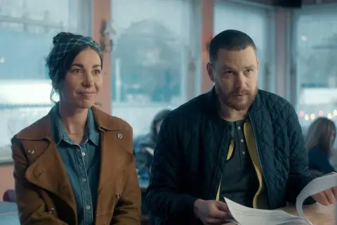 En kvinna med scarf och en man med skägg sitter vid ett bord. Scenen är från en dansk serie på SVT.