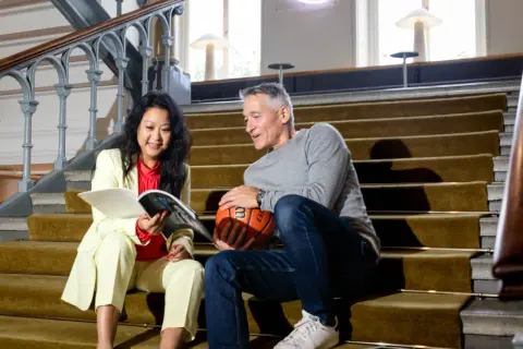 En kvinna och en man sitter på en trappa. Hon läser en tidning, han håller en basketboll. Kanske funderar de på att byta jobb eller inte.