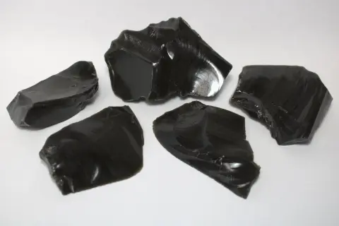 Fünf scharfkantige Stücke glänzenden, schwarzen Obsidiansteins liegen auf weißem Untergrund.