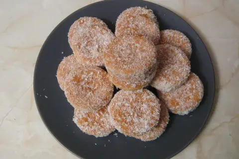 Montón de galletas de coco, doradas y cubiertas de coco rallado, apiladas en un plato oscuro.