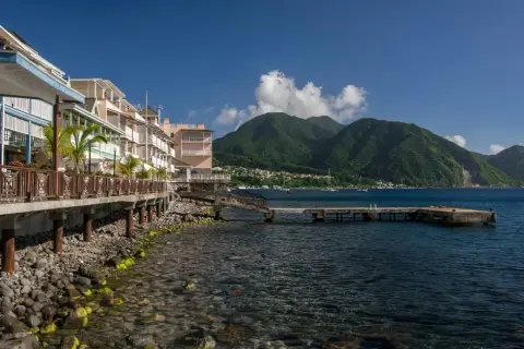 Bâtiments colorés en bord de mer sur l'île Dominique, avec des montagnes verdoyantes en arrière-plan et un quai s'avançant dans l'eau bleue.