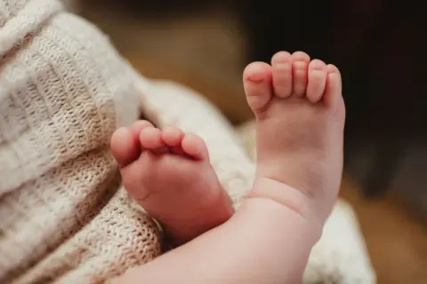 Delikatne, różowe stópki niemowlaka, z widocznymi podwiniętymi palcami u nóg, spoczywają na miękkim, beżowym kocyku.