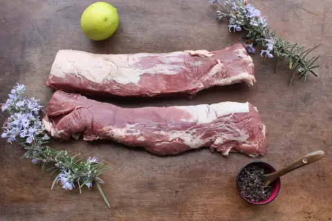 Deux pièces de filet mignon cru sur une planche en bois, accompagnées de romarin fleuri, d'une lime et de baies poivrées.