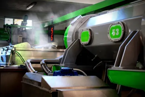 Macchinari industriali in una fabbrica, con tubi e vapore. Si intravede un cartello con "53", forse legato alla produzione di olio di sansa cos'è.