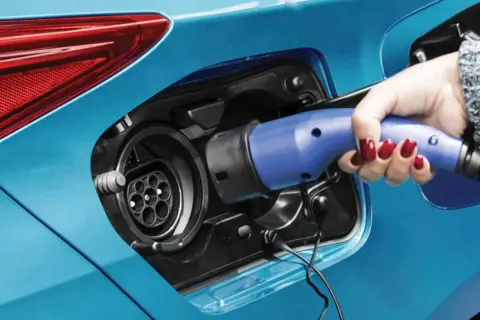 Dłoń z czerwonym lakierem na paznokciach wkłada niebieską wtyczkę do gniazda ładowania samochodu. To przyszłość motoryzacji – hybryda plug-in gotowa do jazdy.