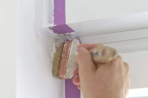 Hand streicht mit Pinsel und Klebeband eine weiße Wand.