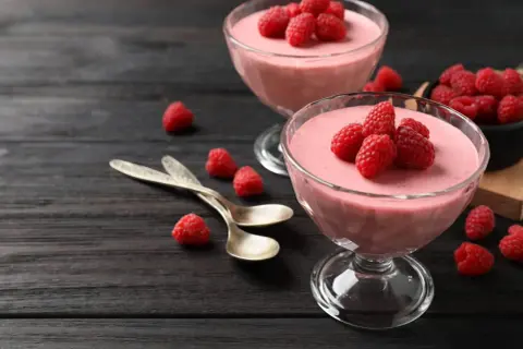 Två glas med hallonmousse, toppade med färska hallon. Två skedar ligger bredvid på ett mörkt träbord.