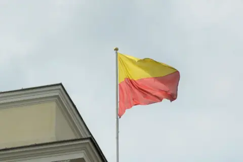 Flaga żółto czerwona powiewa na maszcie obok zabytkowego budynku.
