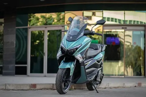 Scooter onex motos color verde menta, con parabrisas alto, estacionada frente a un edificio moderno con puertas de cristal.
