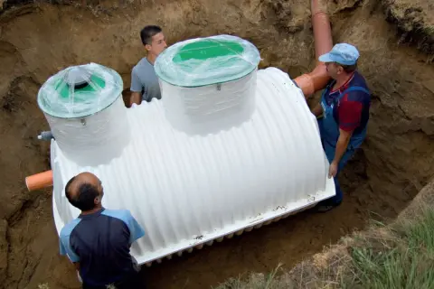 Trzej mężczyźni montują białe szambo betonowe czy plastikowe w wykopie.