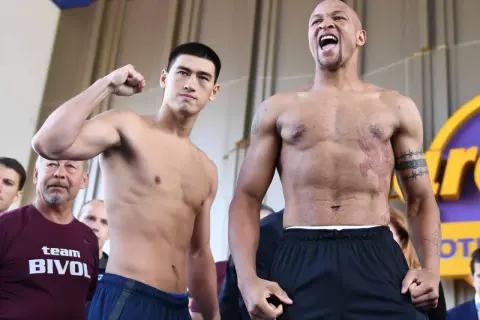 Two boxers face off. One, wearing a "team BIVOL" shirt, flexes his bicep. The other, with tattoos, shouts. Bivol fights in the light heavyweight class.