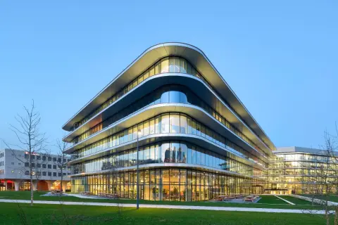 Edificio moderno con **cristales curvos** que reflejan el cielo azul. Fachada ondulada y luminosa, con áreas verdes y árboles alrededor.