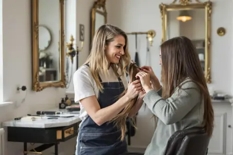 Coiffeuse souriante examine une mèche de cheveux, discutant du type de permanente avec sa cliente assise.