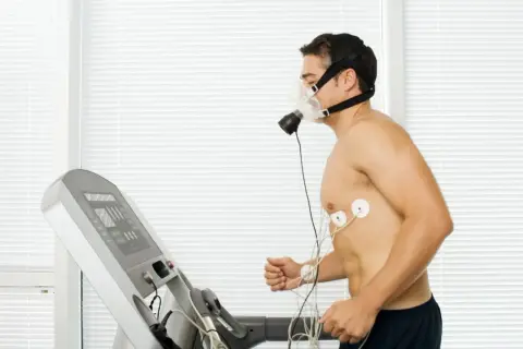 Mężczyzna w masce tlenowej i elektrodach biegnie na bieżni podczas testu wysiłkowego.