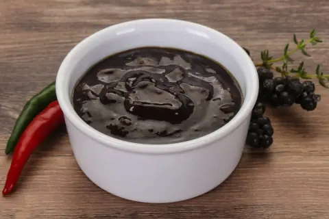 Gęsty, ciemny sos pieprzowy w białej miseczce, obok czerwona i zielona papryczka chili oraz gałązka jeżyn.