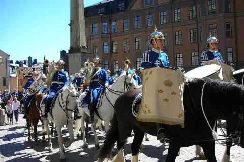 Livgardets dragonmusikkår marscherar i parad. Musiker i blå uniformer spelar på instrument och rider på vita hästar.