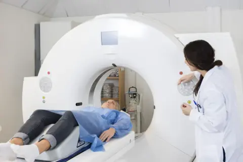 Pacjentka leży na stole tomografu komputerowego, a lekarka przygotowuje badanie. Dowiedz się, ile trwa tomografia komputerowa.