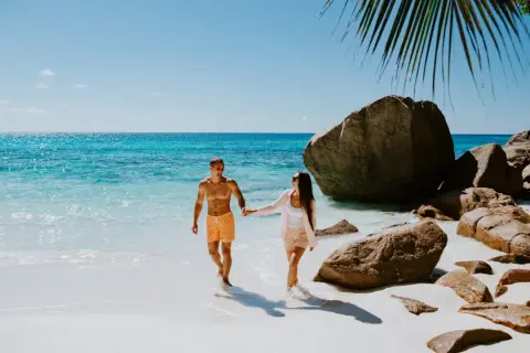 Para idealny czas na podróż na Seszele, para spaceruje po rajskiej plaży z turkusową wodą i skałami.