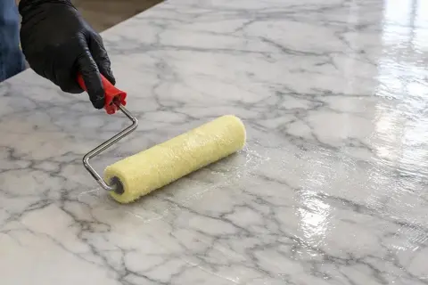 Mano guantata applica resina epossidica per marmo con un rullo, creando una finitura lucida e protettiva.