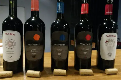 Pięć butelek wina, w tym dwie z etykietą "note nere nero d'avola sicilia doc", stoją na drewnianym stole.