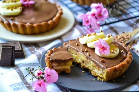 Tarte banane chocolat, garnie de rondelles de banane, de fleurs roses et de noisettes concassées. Un délice gourmand !