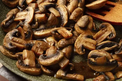 Gebratene Champignons in einer Pfanne, dampfend und glänzend, mit einem hölzernen Kochlöffel.
