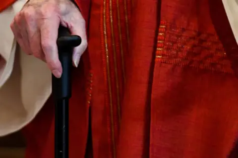 Hand eines Priesters im Ruhestand, der einen Gehstock hält. Er trägt eine rote Soutane mit goldenen Verzierungen.