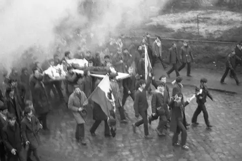 Tłum niesie nosze z ciałem, dym unosi się w powietrzu. Janek Wiśniewski padł, a jego towarzysze idą dalej.