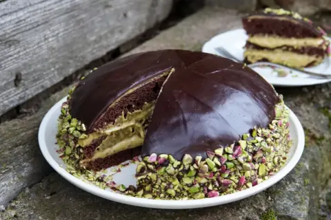 Torta al cioccolato con crema al pistacchio, ricoperta di glassa lucida e granella di pistacchio. Una fetta è già servita.