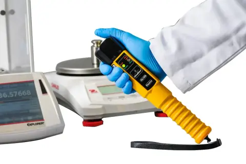 Ręka w niebieskiej rękawiczce trzyma żółty alkomat. W tle waga laboratoryjna.