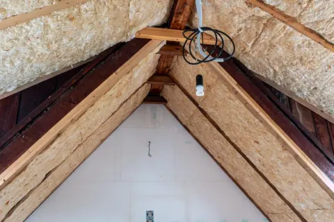 Dämmmaterial wird in einem Dachboden verlegt, um das Haus zu isolieren. Holzbalken und Kabel sind sichtbar.