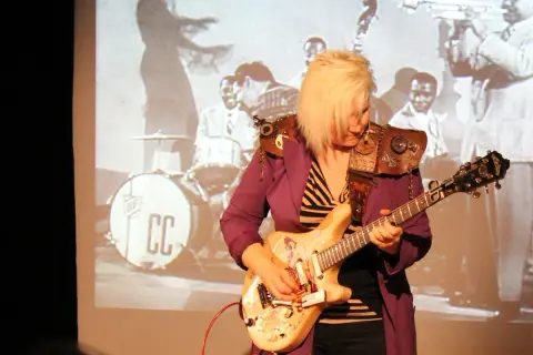 Eine deutsche Gitarristin mit blondem Haar spielt auf einer E-Gitarre vor einer Projektion einer Jazzband.