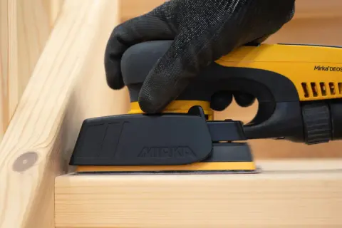 Mano guantata utilizza levigatrice elettrica per carteggiare legno grezzo, preparando la superficie per la finitura.