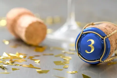 Sektkorken mit blauer Kappe und goldener "3" symbolisiert die Bedeutung des 3. Hochzeitstags. Goldkonfetti und ein weiterer Korken im Hintergrund deuten auf eine Feier hin.