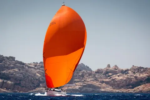 Vela arancione brillante, un gennaker avvolgibile, gonfia al vento su un'imbarcazione a vela che naviga vicino a una costa rocciosa.