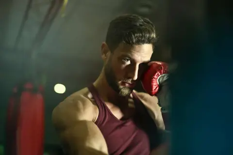 A boxer with a beard and intense blue eyes, wearing a red tank top and a red boxing glove, poses in a gym. He's ready to jump into action.