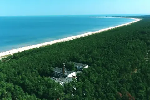 Widok z lotu ptaka na hotel otoczony lasem sosnowym, z plażą i morzem w tle.