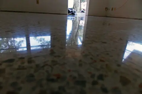 Reflejos en suelo de terrazo pulido, mostrando la entrada a una casa con bolsas de basura y un árbol. Se observa humedad en suelo de terrazo.